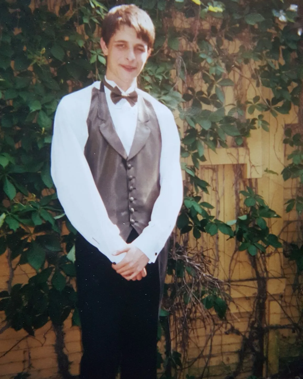 Teenage Jake in a vest and bow tie, standing in front of a fence covered in vines