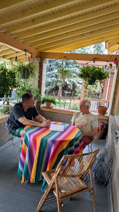 Jake and a neighbor talking across a rainbow tablecloth at a porch forum, hanging plants overhead
